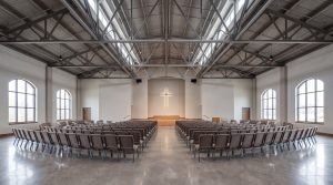 "Interior of a modern church with rows of chairs facing a raised platform, large arched windows, and exposed steel trusses on the ceiling, featuring a lit cross on the wall."