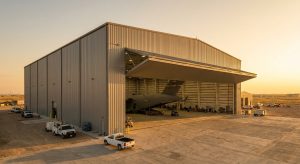 A large metal airplane hangar at sunset with an open door, showing a military cargo plane inside and several service vehicles parked outside, illustrating future-proof hangar design for accommodating wide-body aircraft.