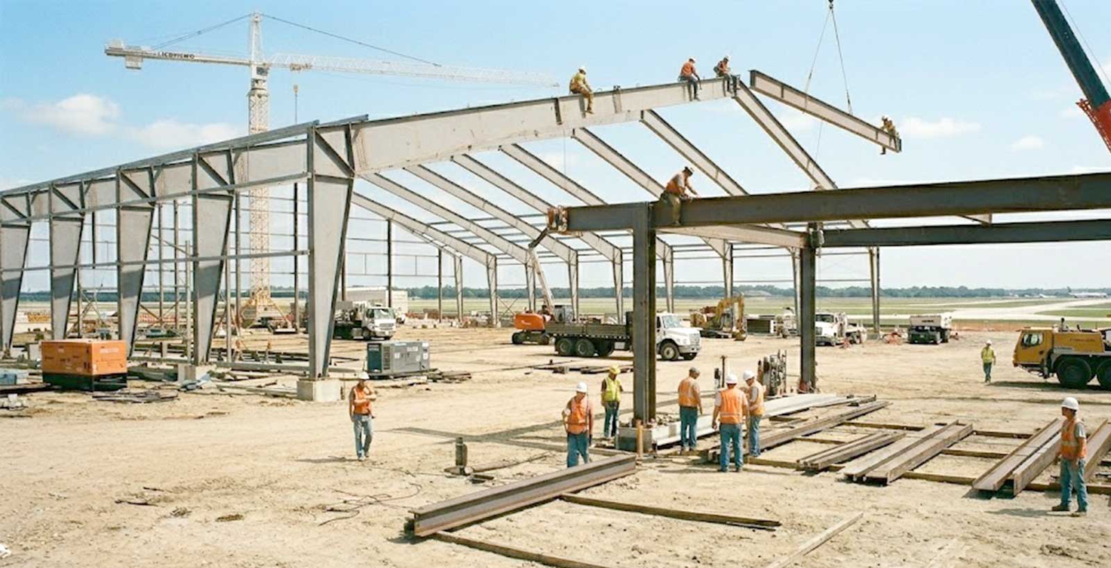 Aircraft hangar construction site with prefabricated steel frames being lifted and assembled by cranes and workers.