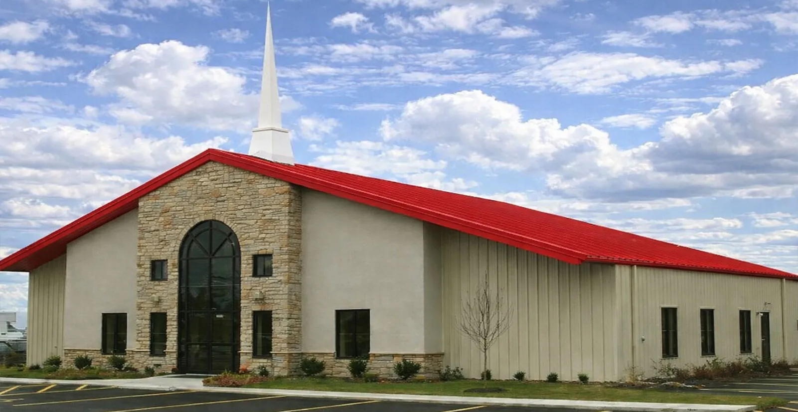 Church building architecture with red metal roof, stone front façade, arched glass entrance, beige wall panels, and tall white steeple for worship projects.