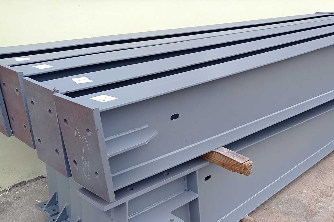 Fabricated steel structure warehouse frame members with welded stiffeners, bolted end plates, and gray protective coating.