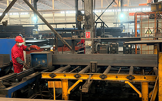 Worker assembling H-section steel beam components on a build-up line in the structural steel fabrication workshop.
