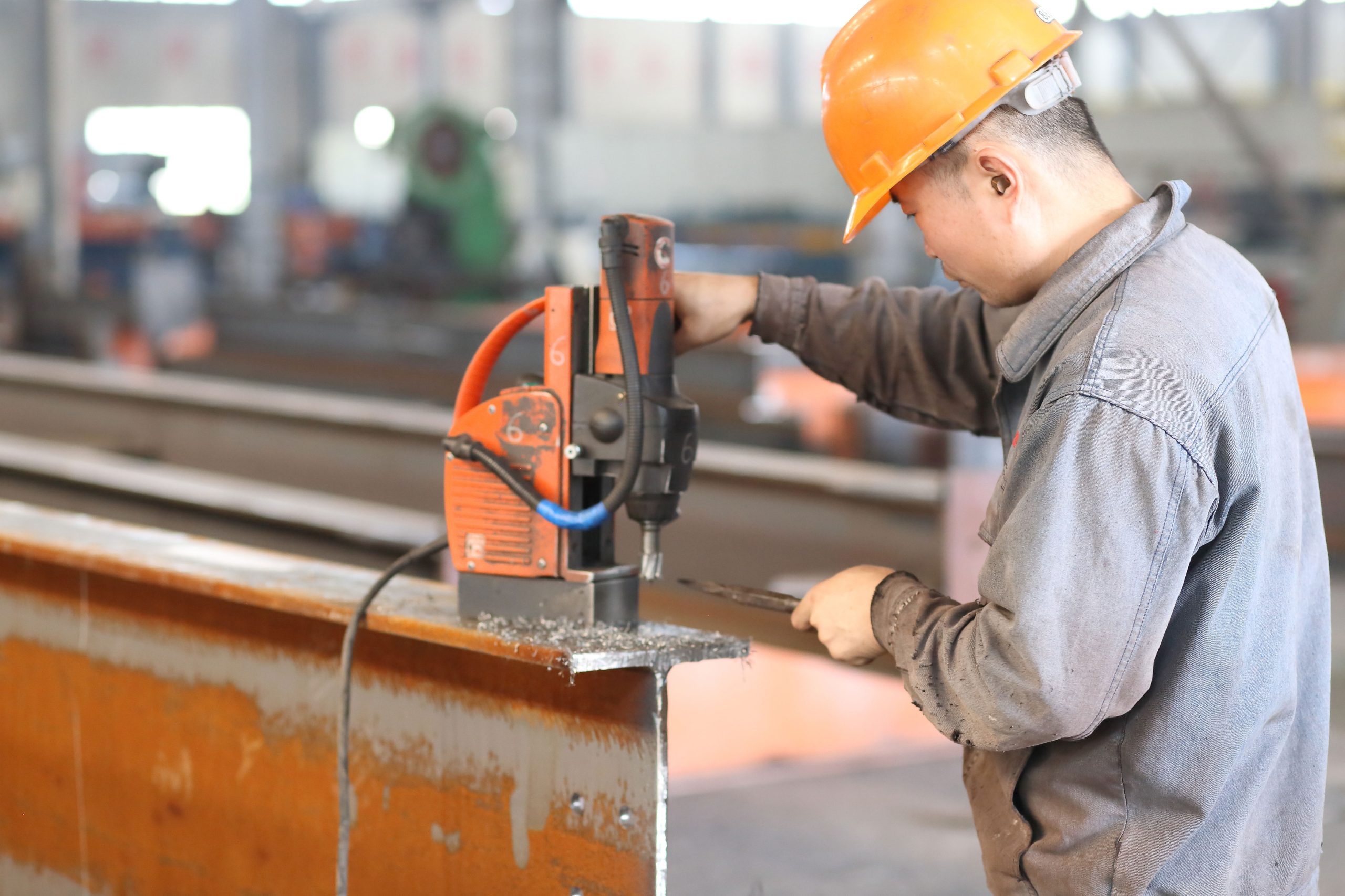 Worker drilling connection holes in an H-section steel beam with a magnetic drill inside a steel fabrication workshop.