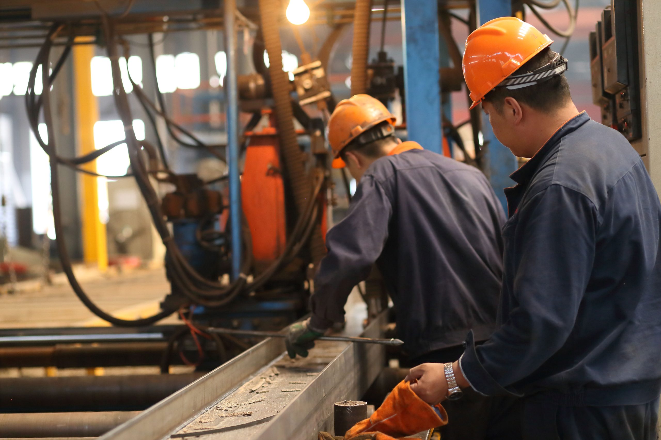 Workers removing welding slag and checking the seam on H-section steel beams during the steel structure building process.