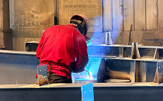 Worker fitting and aligning steel plates on an H-section beam in the structural steel fabrication workshop.