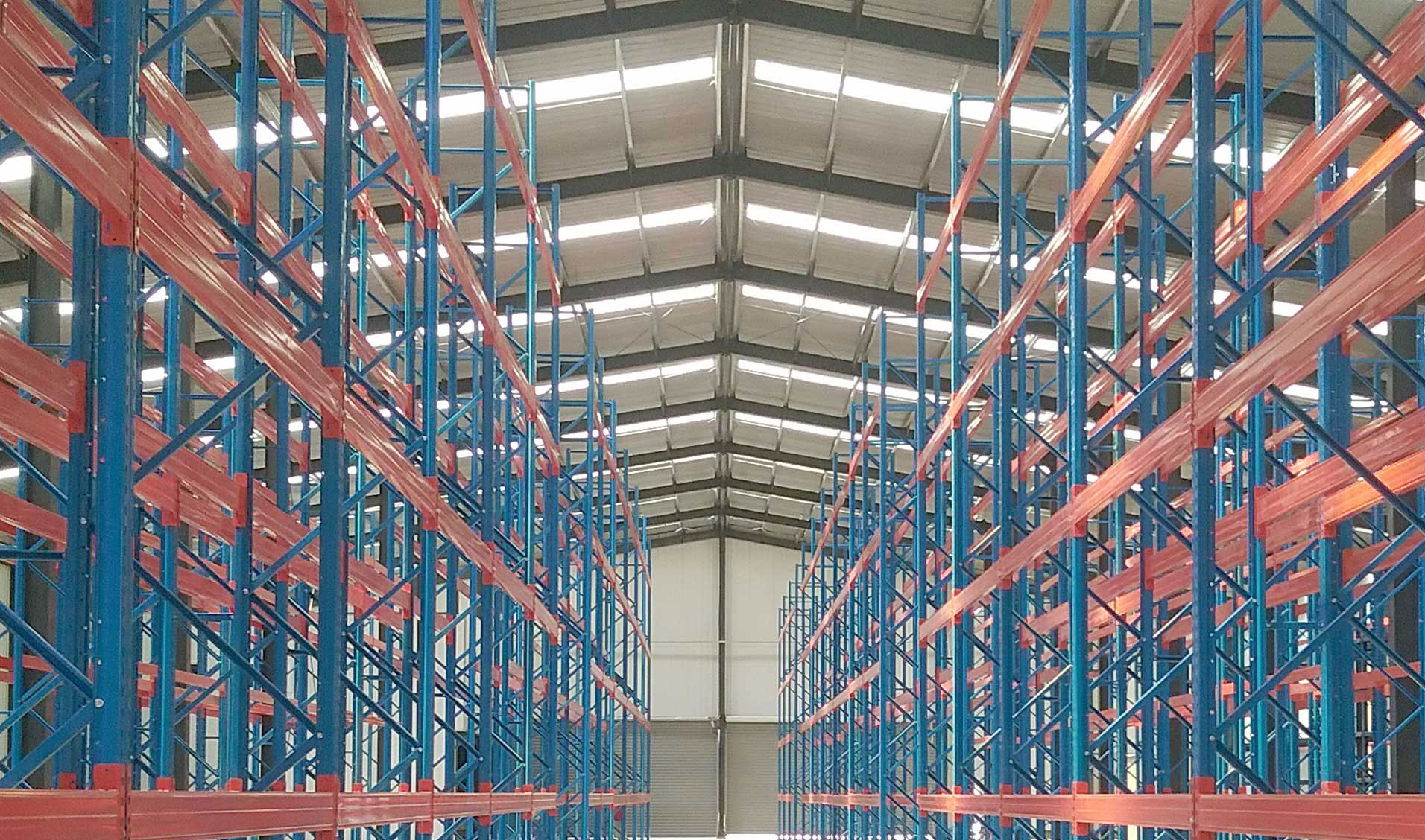 Steel structure warehouse interior with high-bay pallet racks, roof skylights, and a central storage aisle.