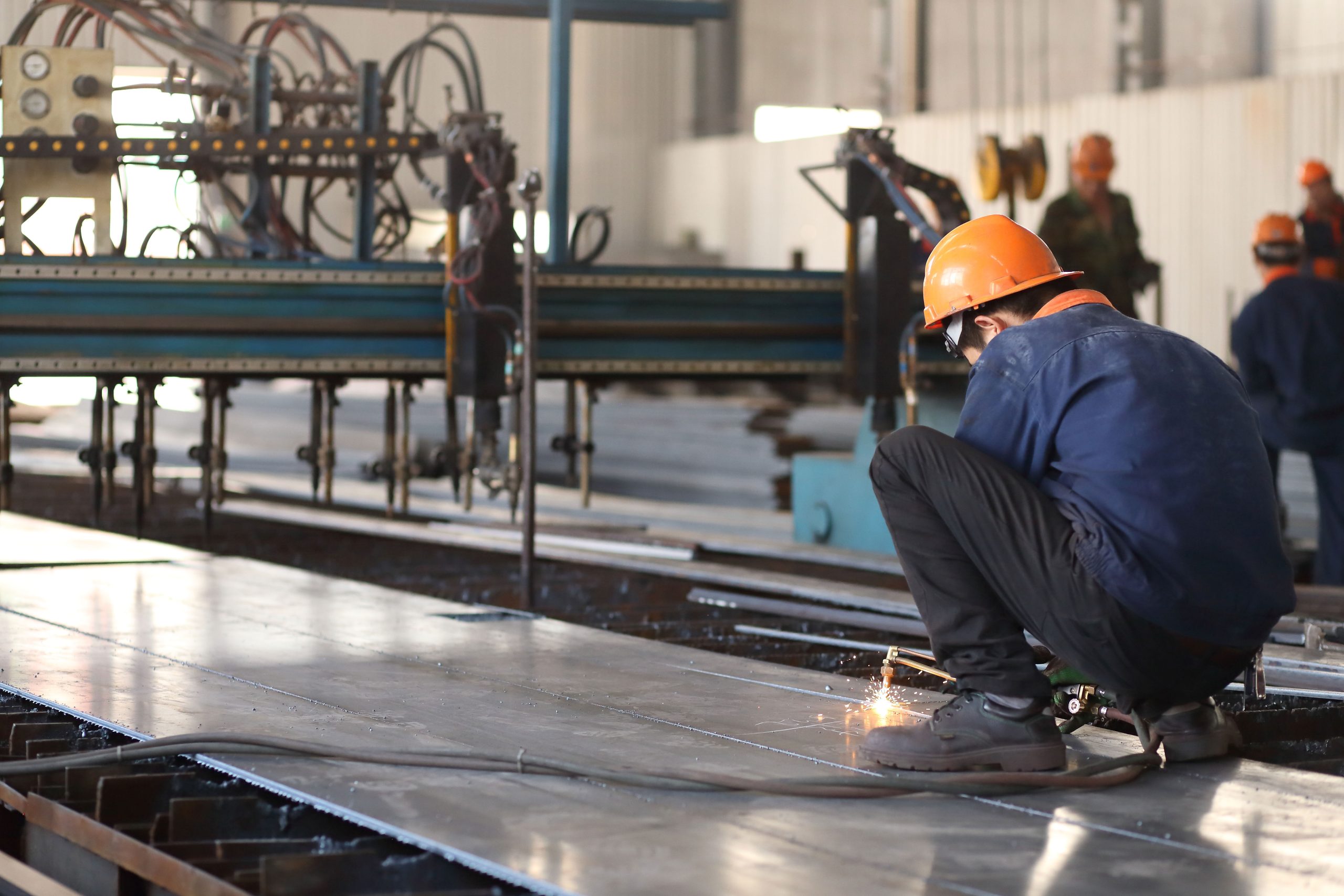 Worker manually cutting steel plate for building structure components beside a multi-head cutting line in the fabrication workshop.
