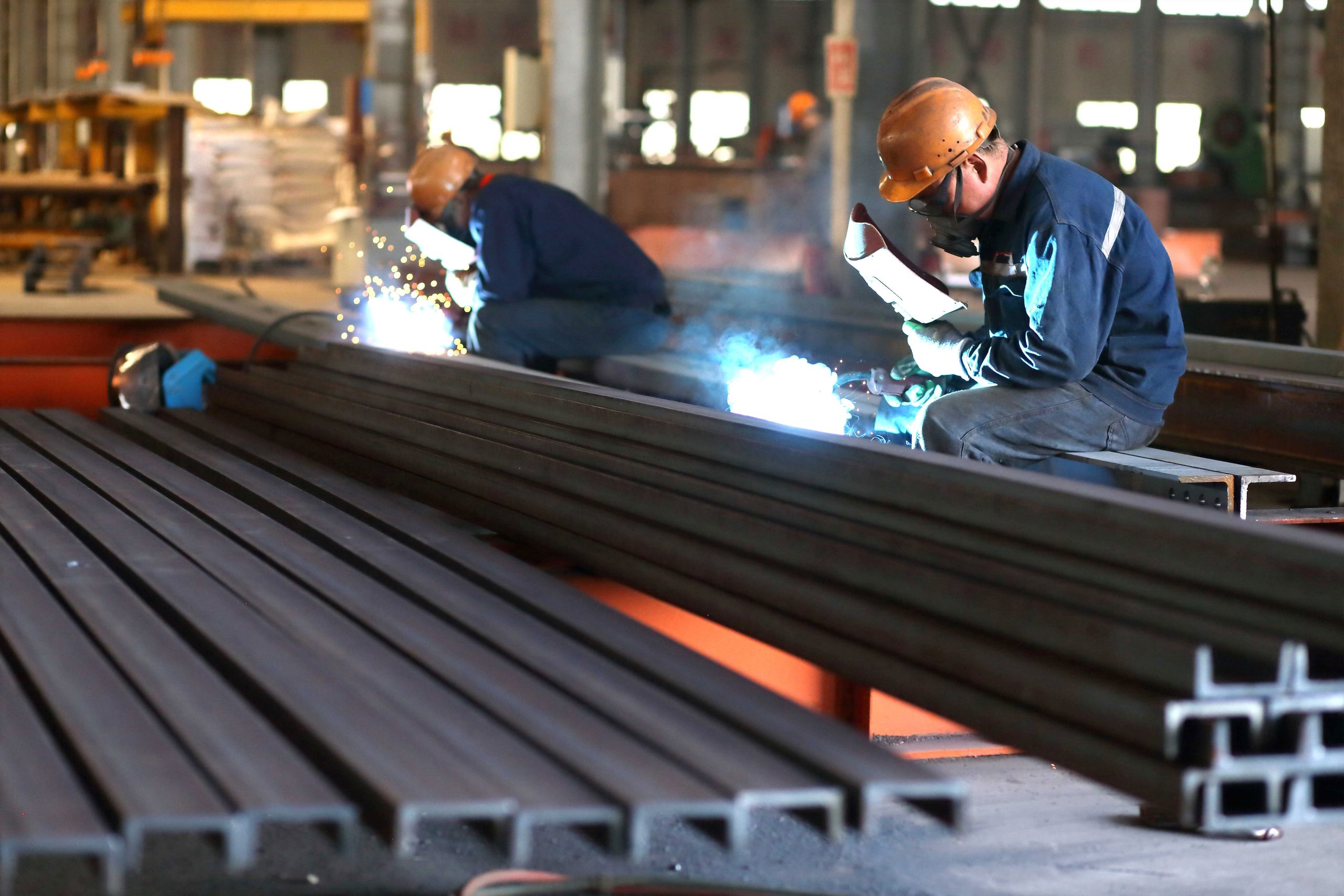 Workers manually welding steel structure building components beside stacked rectangular steel sections in a fabrication workshop.