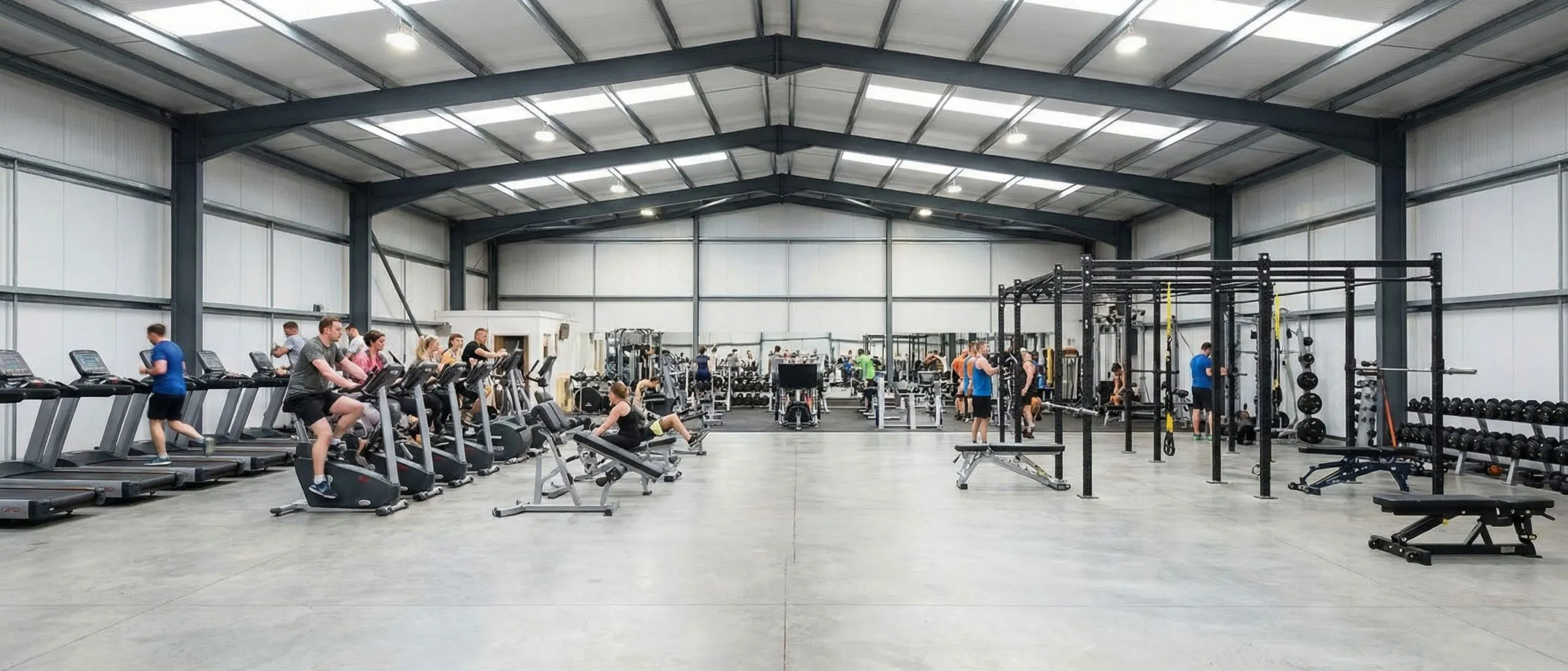 Large open-span steel gym interior with cardio machines, free weights, training rigs and clear circulation space.