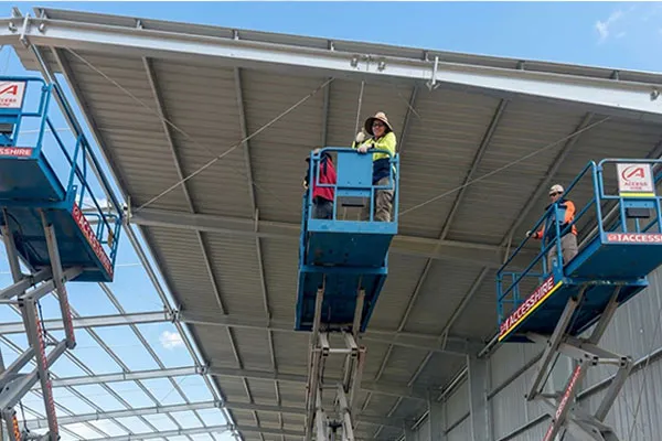 Oceania steel structure warehouse project with workers installing metal roof panels using aerial lifts during on-site steel building construction.