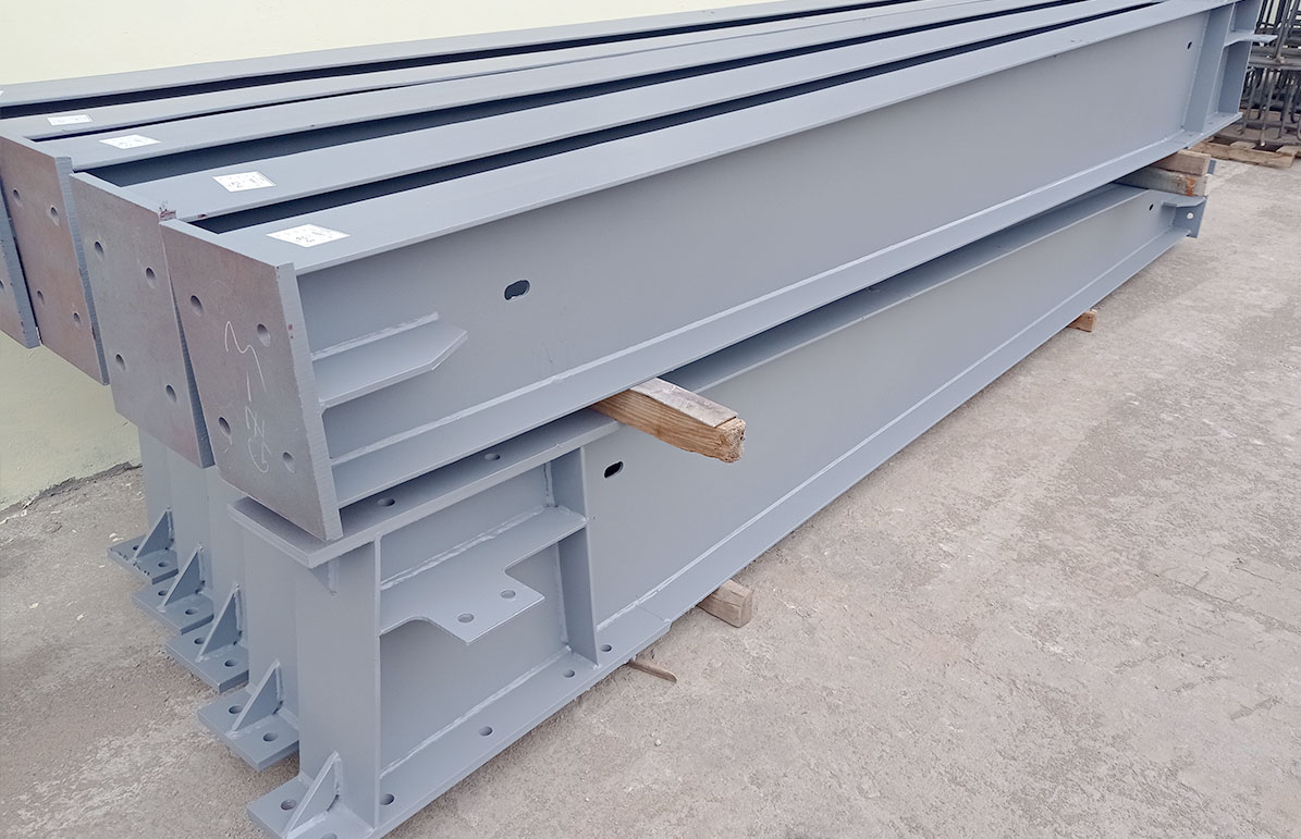 Painted H-section steel beams with drilled end plates, welded stiffeners, and mounting brackets stacked for building assembly.