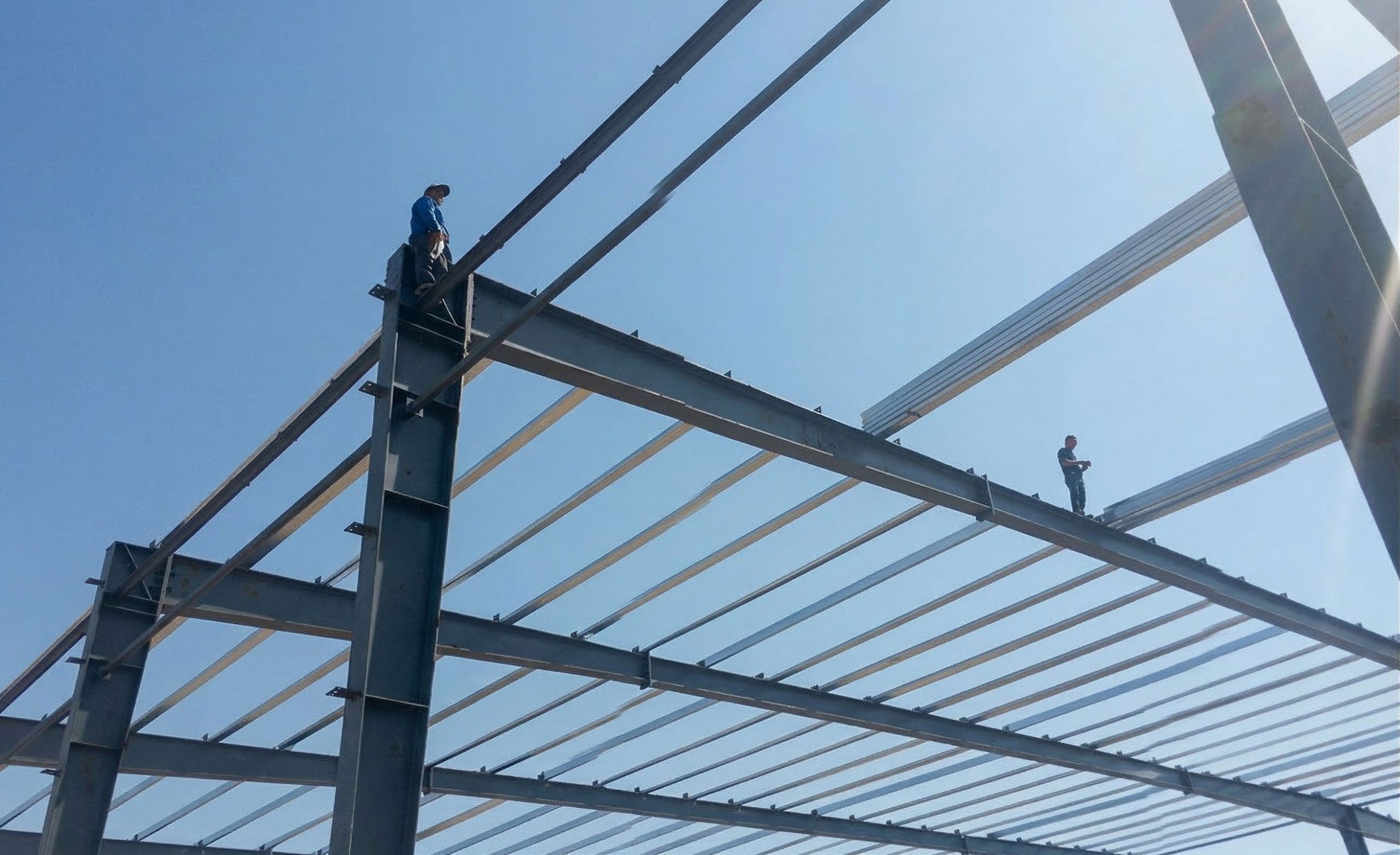 Workers installing roof purlins across steel rafters during secondary framing of a steel structure building.