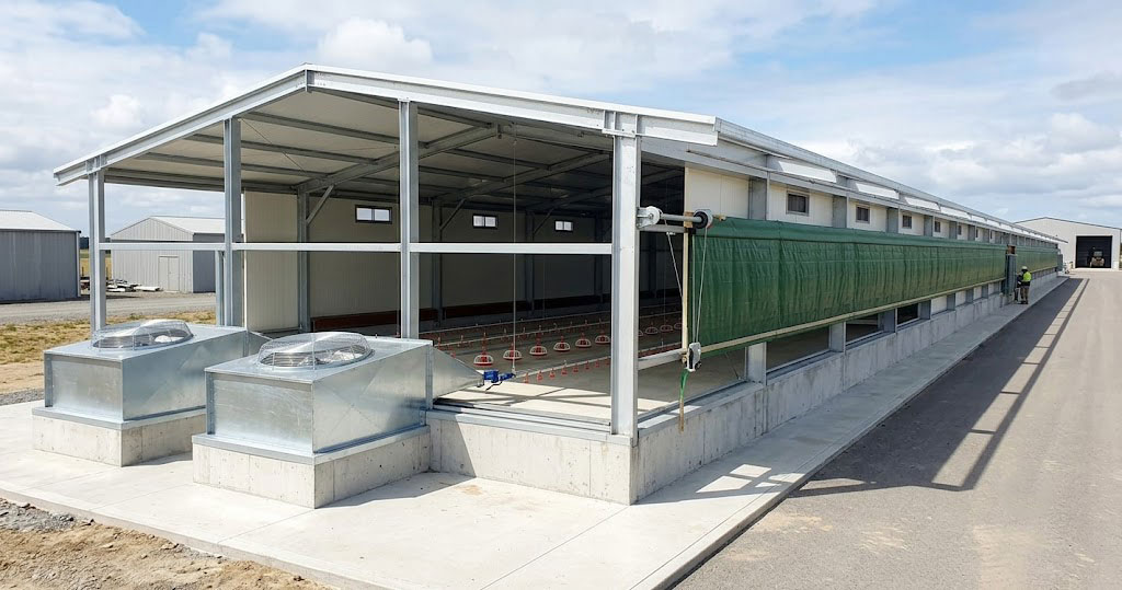 Exterior of a semi-enclosed chicken house with roll-up side curtains, steel framing and ventilation equipment.