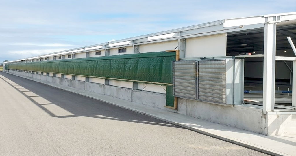 Exterior of a semi-enclosed chicken house with green roll-up side curtains for adjustable natural ventilation.