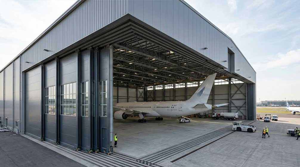 Sliding aircraft hangar doors open across a wide steel hangar entrance with a large passenger aircraft positioned inside.