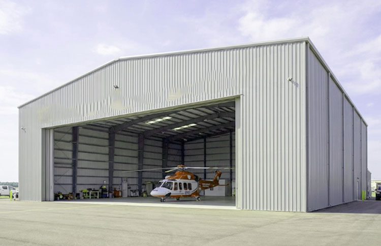 Steel aircraft hangar with a wide open entrance and a helicopter parked inside on an airport apron.
