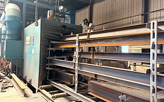 Structural steel components undergoing sandblasting for surface preparation in an industrial steel fabrication workshop.
