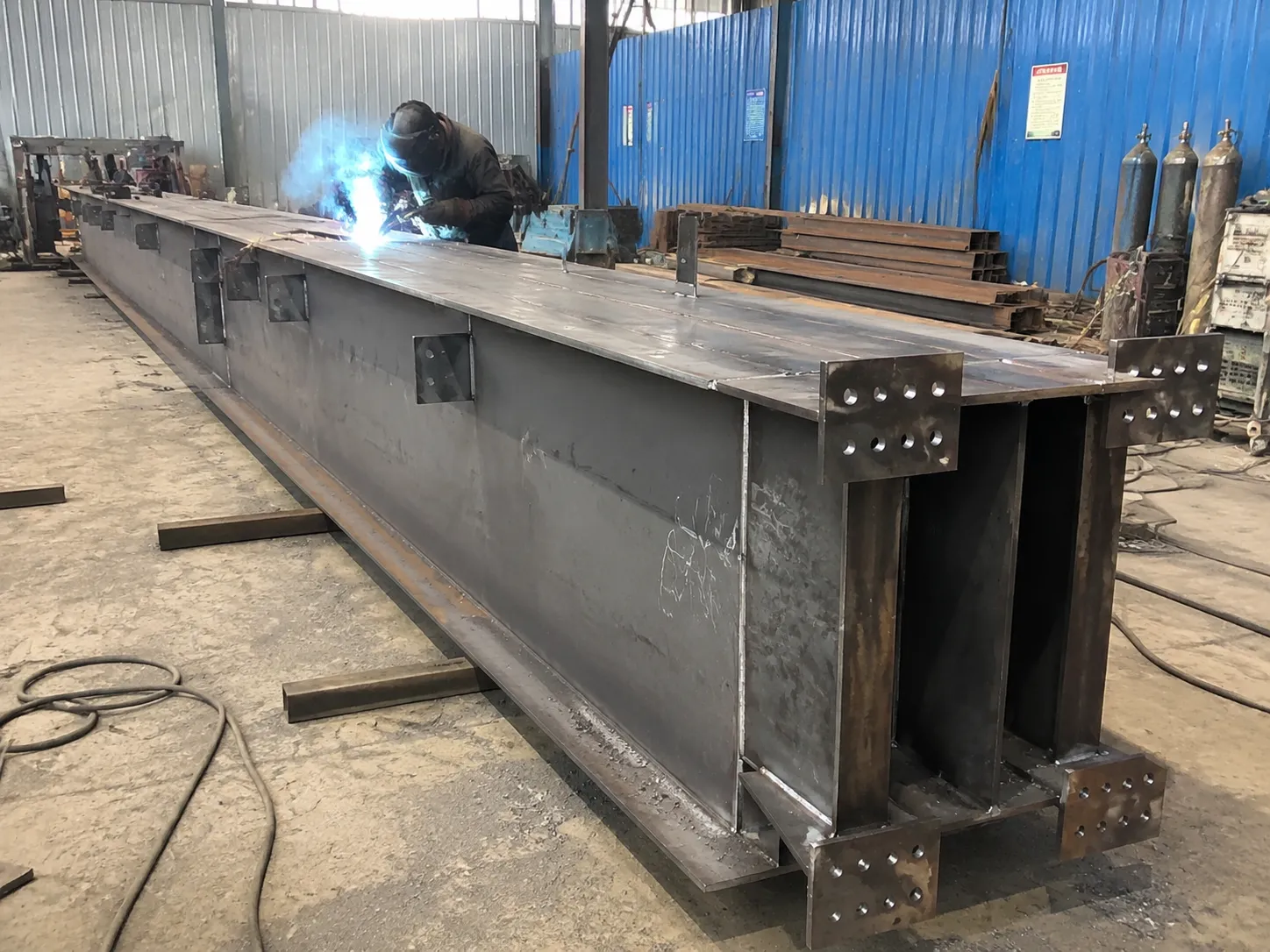 Worker welding H-section steel members in the fabrication workshop for the 4,320 m² Bolivia warehouse project.