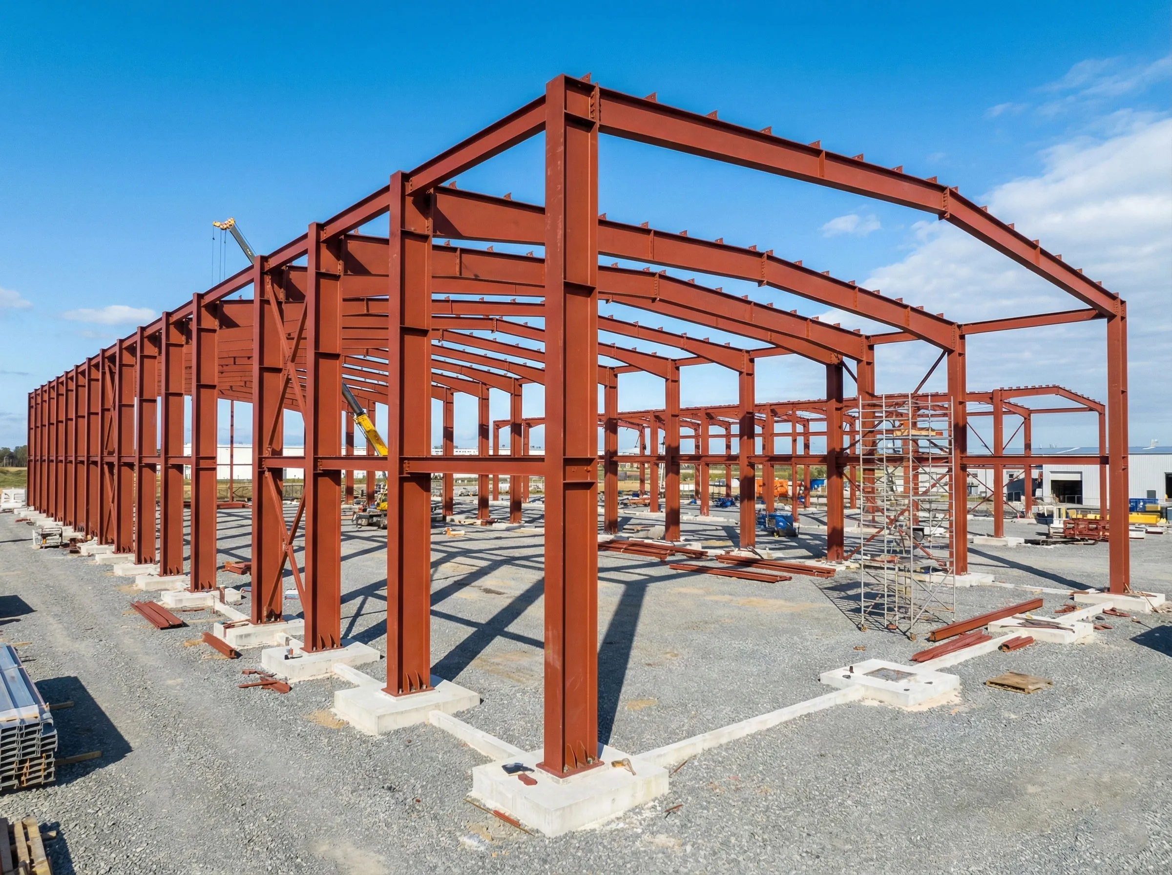 Steel frame workshop structure under installation with portal steel columns, roof beams, crane lifting work, and concrete foundations.