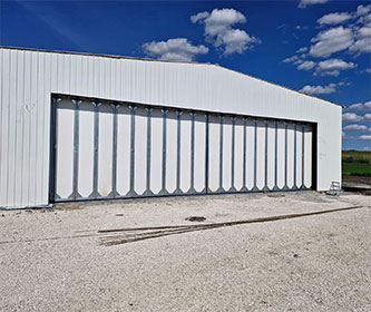 Steel structure aircraft hangar exterior with a wide multi-panel door, metal wall cladding, and an open access apron.