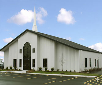 Steel structure church building with white metal wall cladding, a front steeple, arched entrance glazing, and parking area.