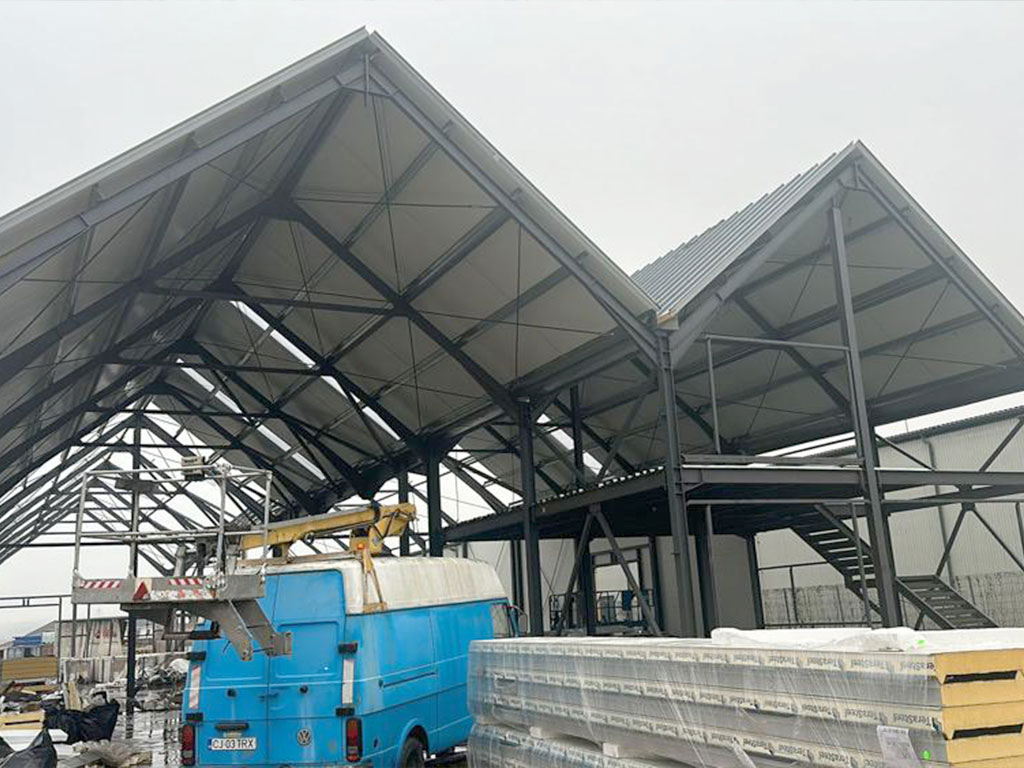 Steel structure industrial building project in Romania with gable roof frames, steel bracing, a raised platform, and roof panel installation.