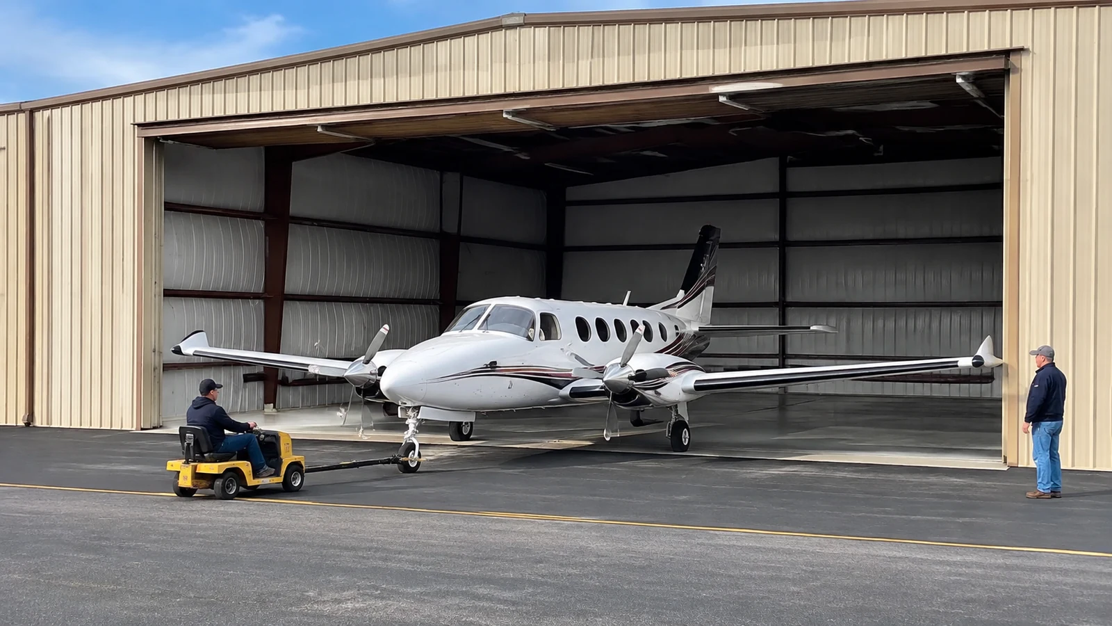 A tug moves a twin-engine aircraft through a wide hangar opening while a spotter monitors clearance.