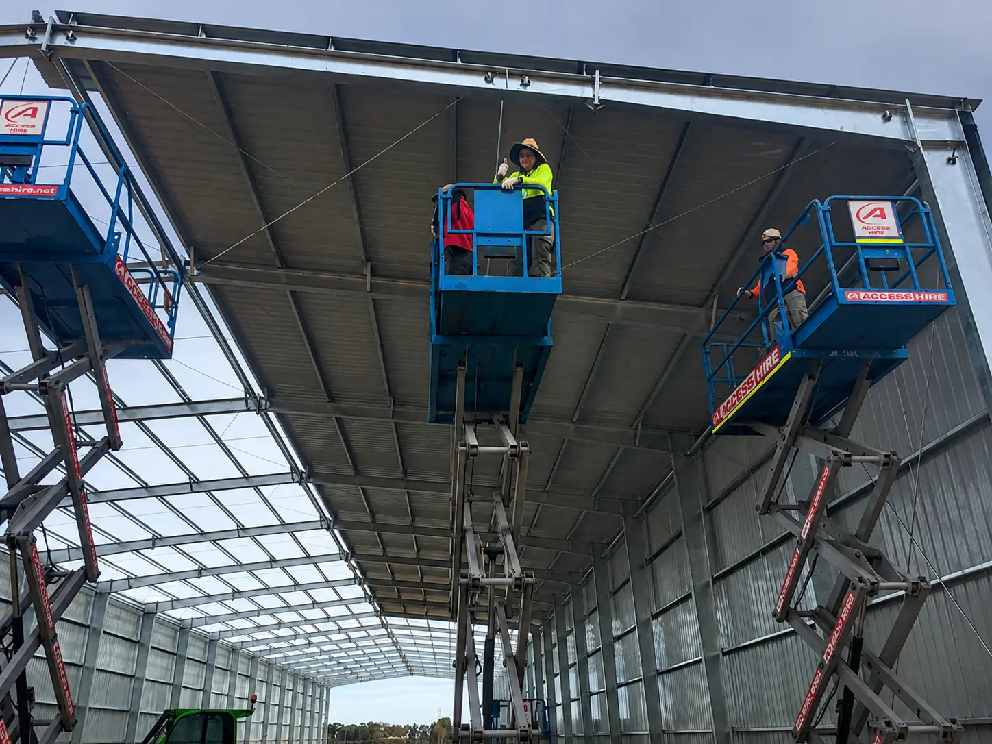 Workers install roof panels on an agricultural steel warehouse under construction in Australia using scissor lifts.