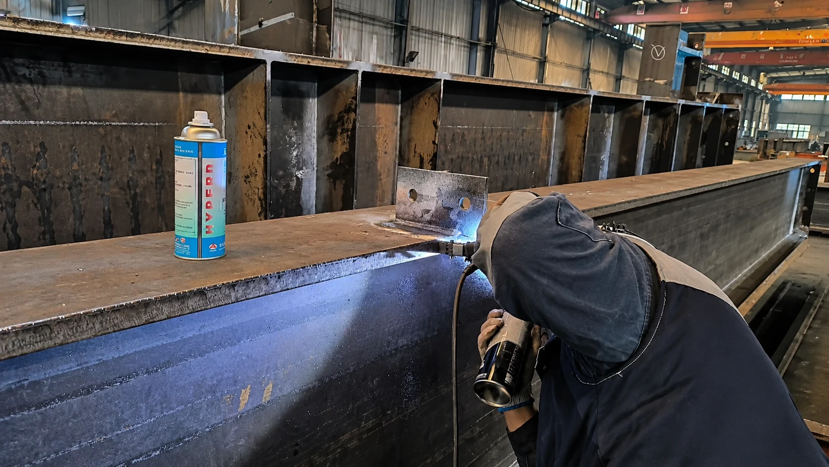 QC inspector checking H-beam welding quality during steel structure fabrication on the factory production floor.
