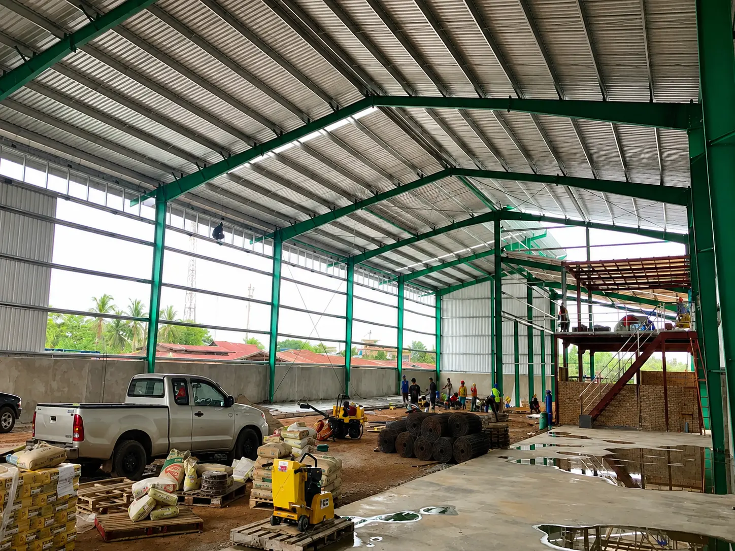 Interior of a Thailand steel warehouse under construction with portal frames, roof cladding, workers and site materials.