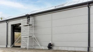 Workers install insulated warehouse wall panels and gutters from a guarded scaffold tower