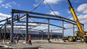 Crew erects a steel workshop portal frame with crane support and temporary bracing in place.